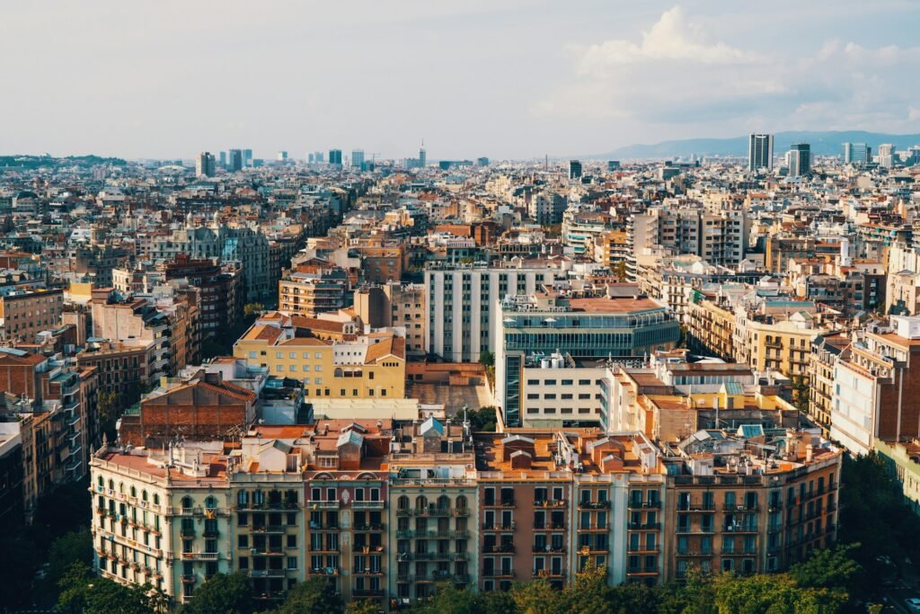 An elevated, sprawling cityscape view of Barcelona, Spain, showing the dense urban architecture and skyline on a partly cloudy day.