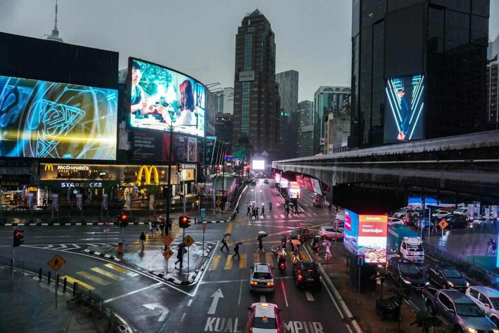 A busy street intersection in Kuala Lumpur, Malaysia, on a rainy day, with large digital billboards and modern skyscrapers.