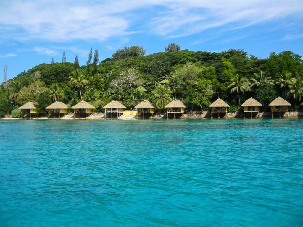 A row of traditional thatched-roof bungalows on stilts along the shore of a vibrant turquoise bay in Vanuatu.
